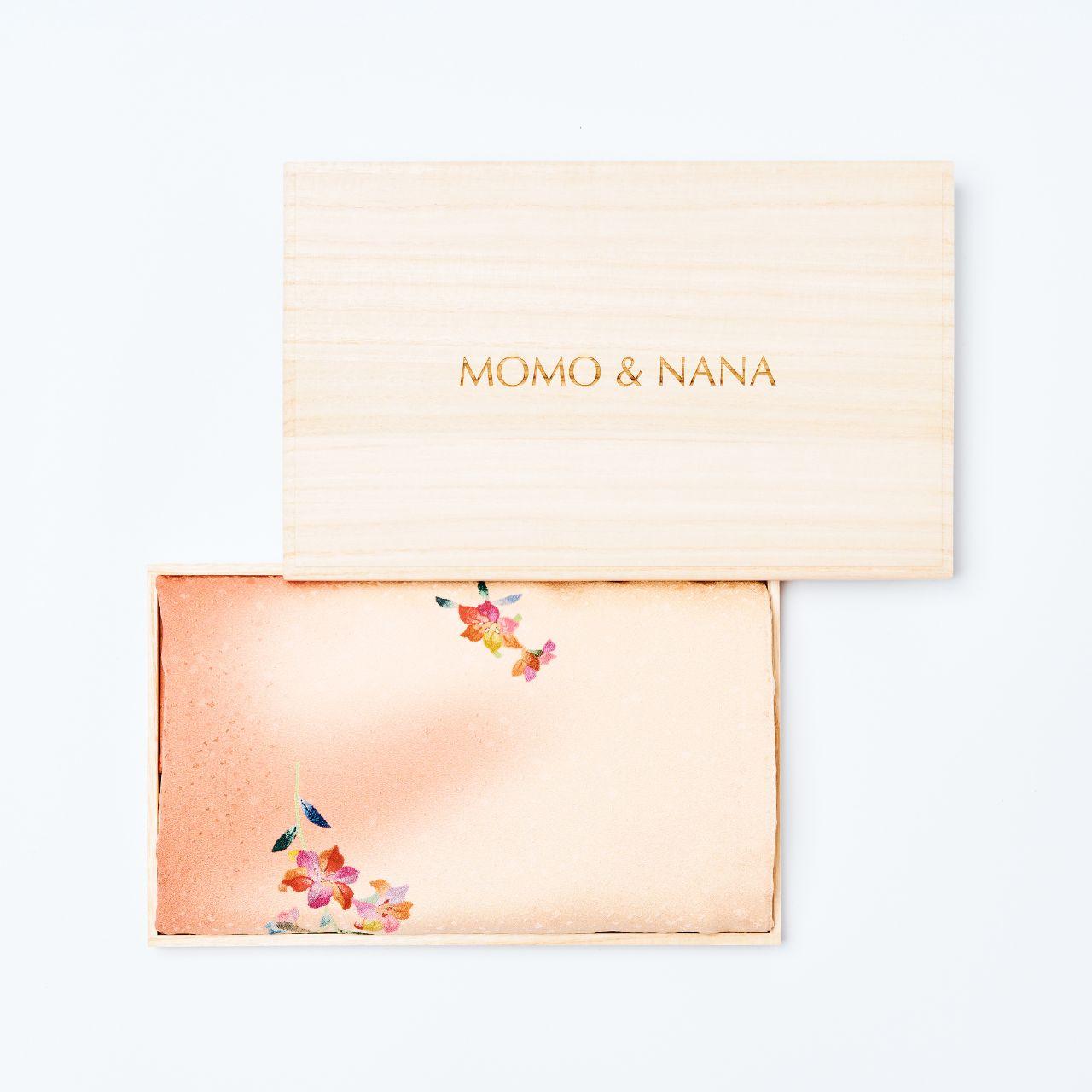 A luxury wooden gift box with the "MOMO & NANA" logo engraved, containing a folded pale crimson and cream Japanese silk fabric with floral embroidery.