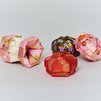 A collection of traditional Japanese fabric boxes in various colors including pink, red, and black gold brocade silk.